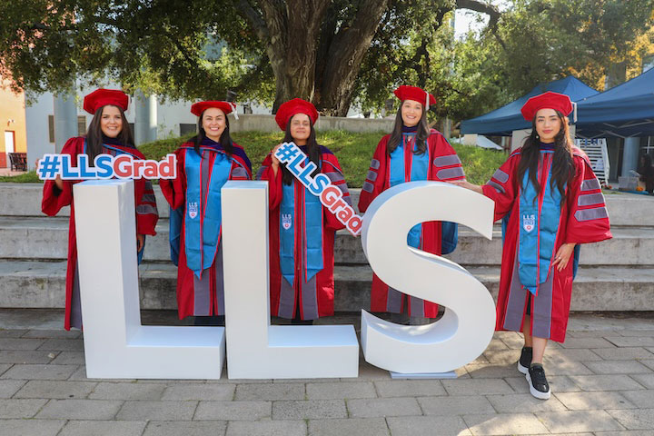 Graduates in cap and gown holding hashtag LLSGrad signs while standing near a large standee of the letters LLS