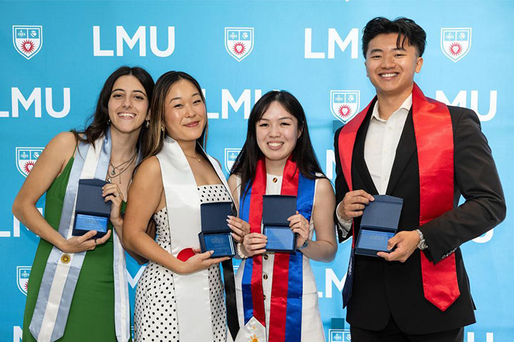 Several graduates holding awards and standing in front of an LMU branded wall